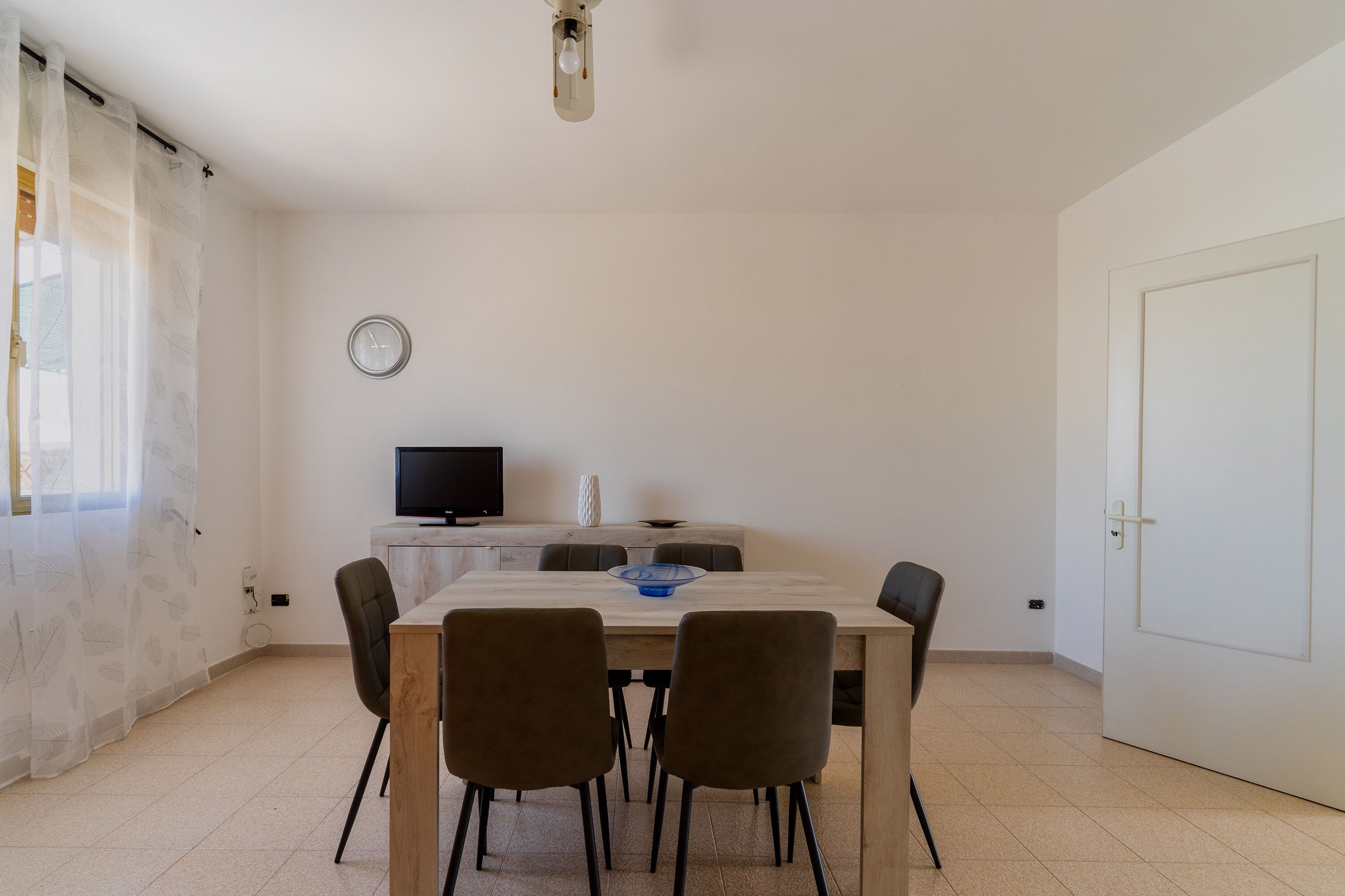 Un comedor con una mesa de madera, cuatro sillas, un televisor sobre un mueble bajo y un reloj en la pared.