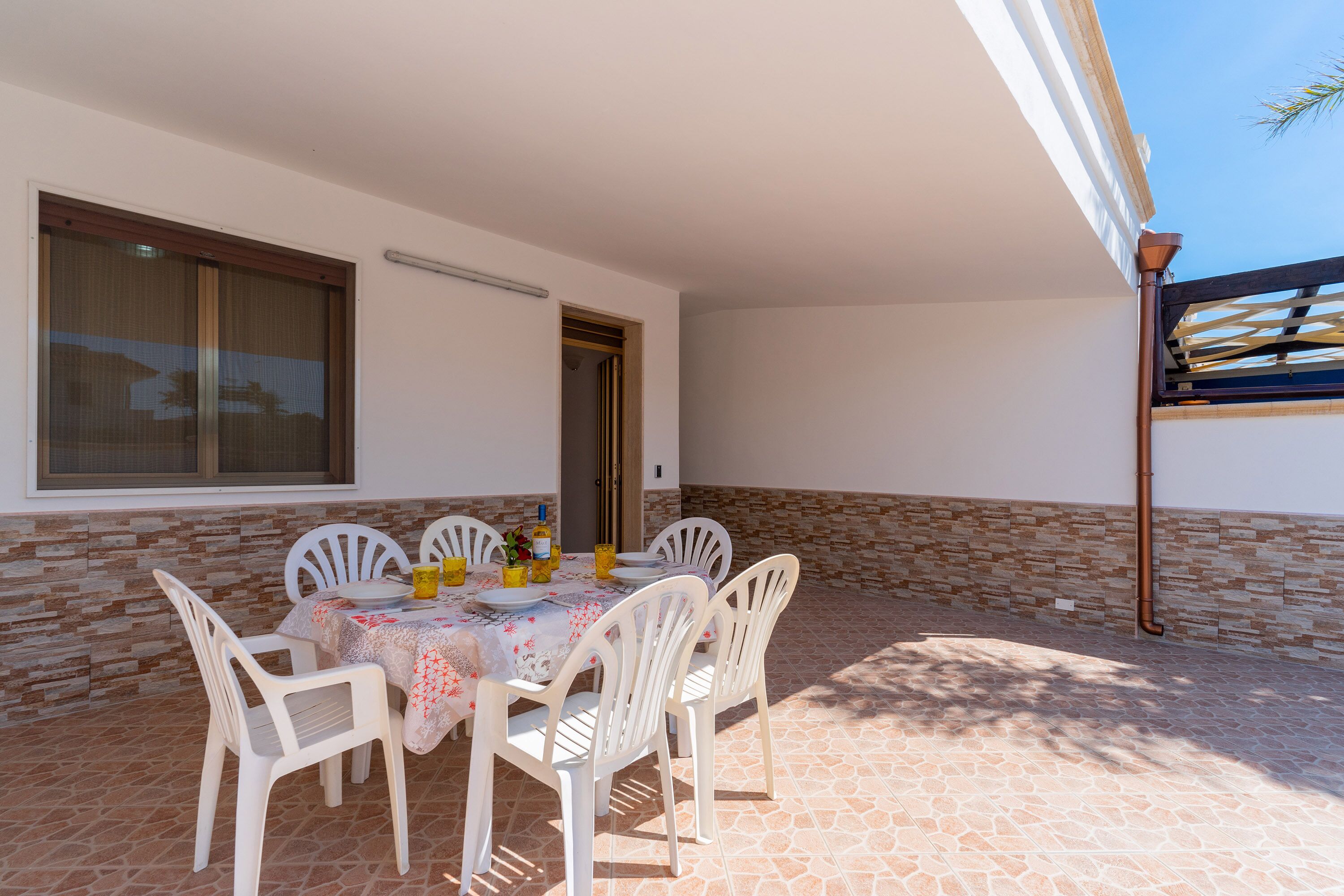 An outdoor patio with a table set for four, surrounded by white plastic chairs, and a stone-tiled floor.