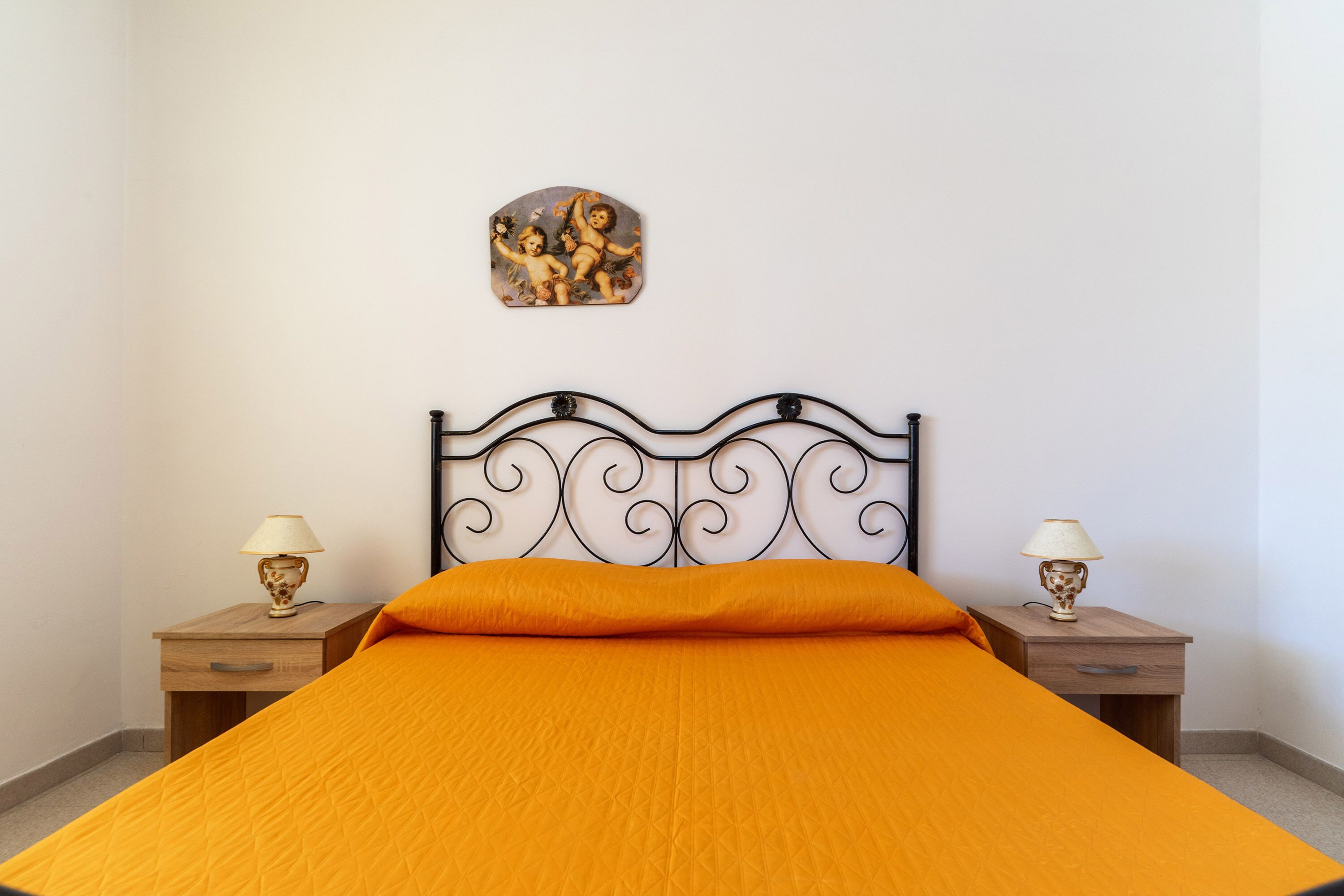 A neatly made bed with an orange bedspread and a decorative headboard. There are two bedside tables with lamps and a small painting above the headboard.