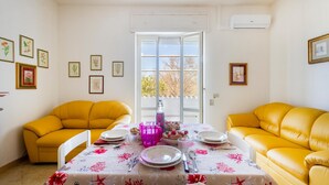 A dining area with a table set for two, surrounded by yellow sofas, a window with a view, and framed botanical prints on the walls. - 2922 Villetta Marelia by Barbarhouse (Porto Cesareo)