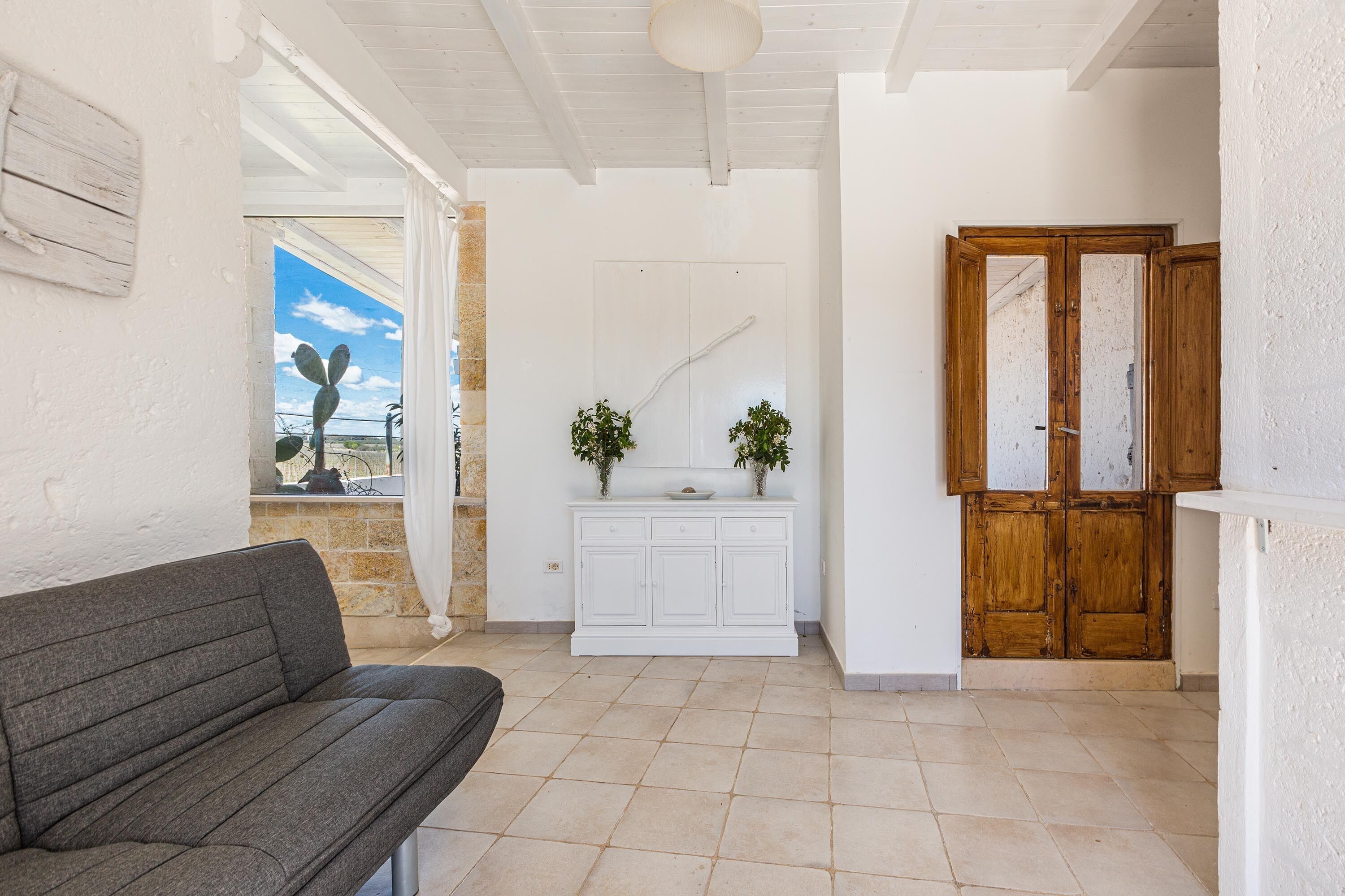 Quarto com sofá cinzento, um armário branco com plantas, porta de madeira e vista do céu pela janela.