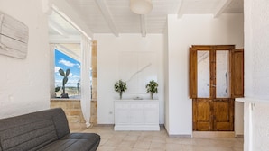 A room with a grey sofa, a white cabinet with plants, a wooden door, and a view of the sky through a window. - 3003 Masseria Surani Grande- Appartamento Sinistra by Barbarhouse (Manduria)