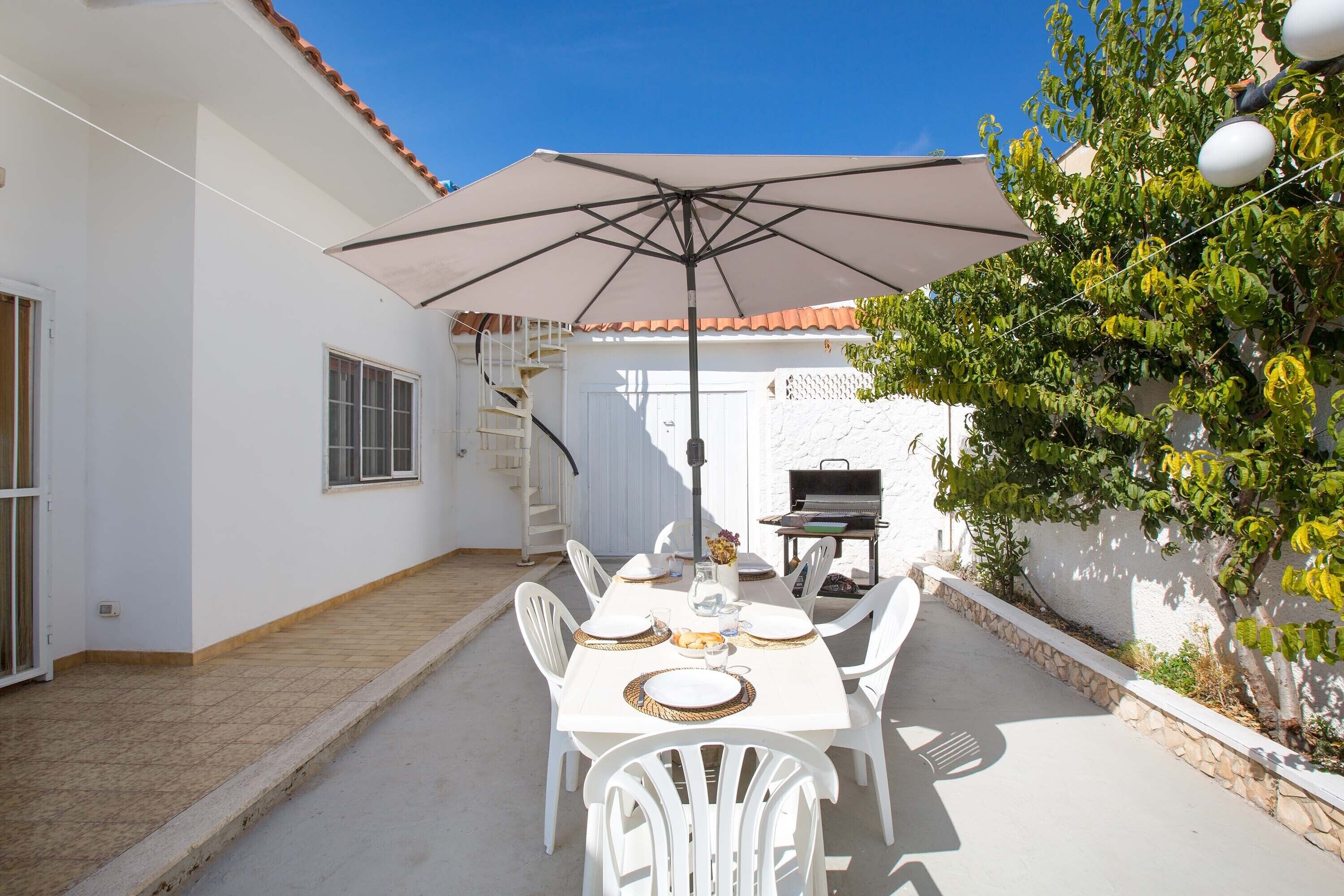 A patio with a table set for four, a grill, and a white wall with a tree.