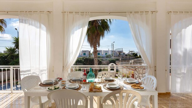 A dining area with a table set for four, white chairs, and a view of buildings and palm trees through sheer curtains. - 2725 Villa Beatrice by Barbarhouse (Manduria)