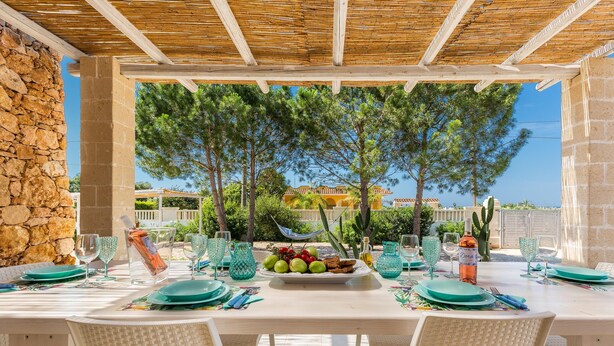An outdoor dining area with a stone wall, wicker chairs, and a table set for two with a fruit bowl and a bottle of wine.