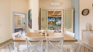 A dining area with a wooden table and white chairs, an open door leading to a balcony, and a clock on the wall.