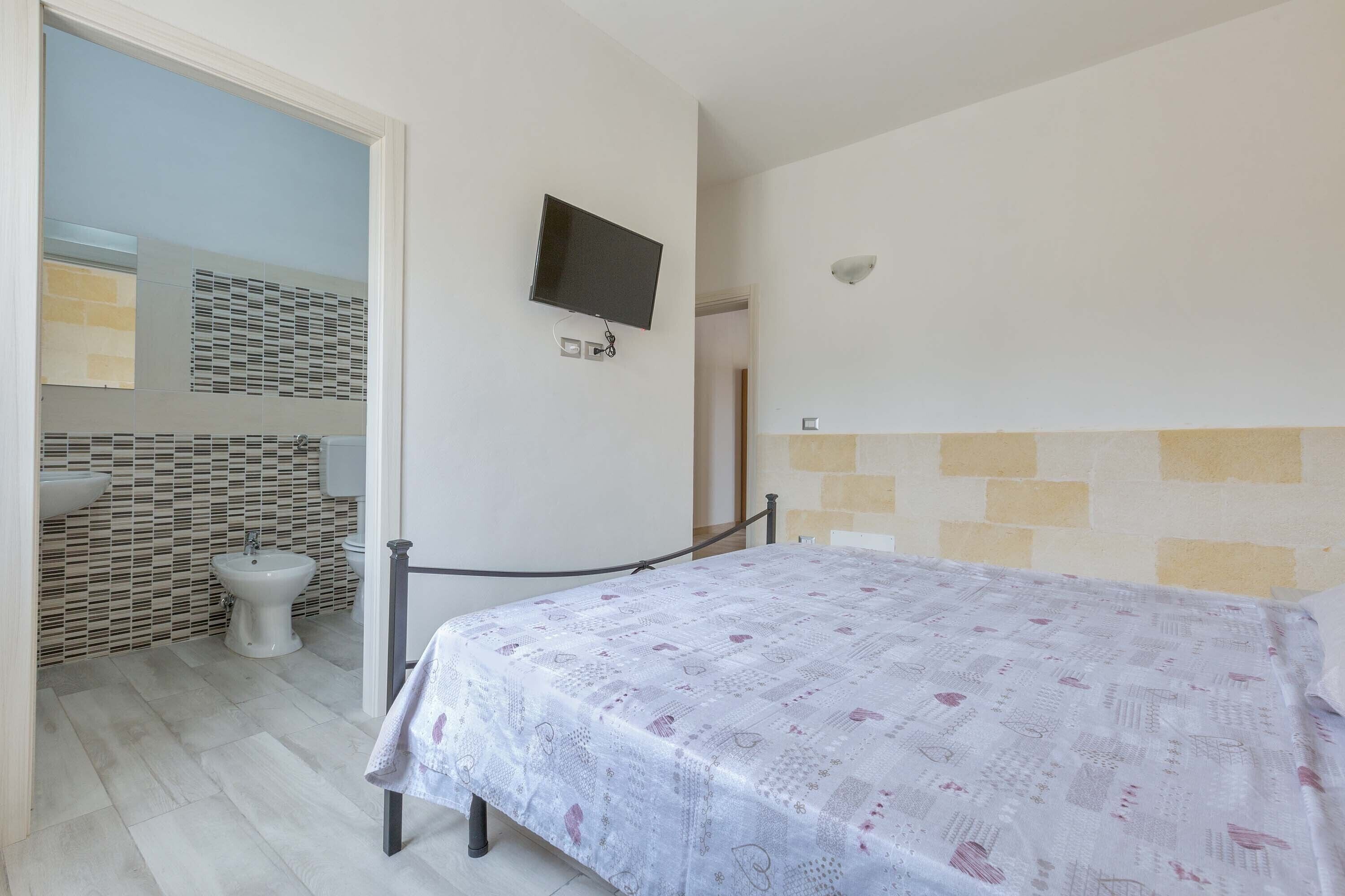 A bedroom with a bed, a TV mounted on the wall, a bathroom with a tiled backsplash, and a sink.
