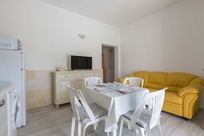 A compact living space with a dining table set for four, a yellow sofa, a washing machine, and a television on a cabinet. - 3064 Villa La Fenice by Barbarhouse (Avetrana)