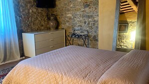 Een slaapkamer met een groot bed, een dressoir, een televisie bevestigd aan een stenen muur en een spiegel die een deel van de kamer weerkaatst.