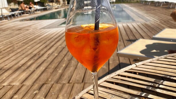 Een glas sinaasappeldrank op een houten terras, met een zwembad en ligbedden op de achtergrond.