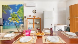 A dining area with a table set for two, a painting of a beach scene, a washing machine, and a refrigerator. - 3028 Villino Dolceluna by Barbarhouse (Porto Cesareo)