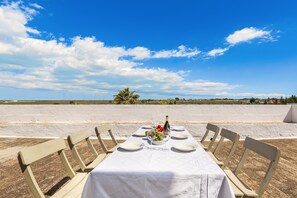 A table set for two with a white tablecloth, two chairs, and a bottle of champagne. The table is set on a sandy beach with a clear blue sky and a palm tree in the background.