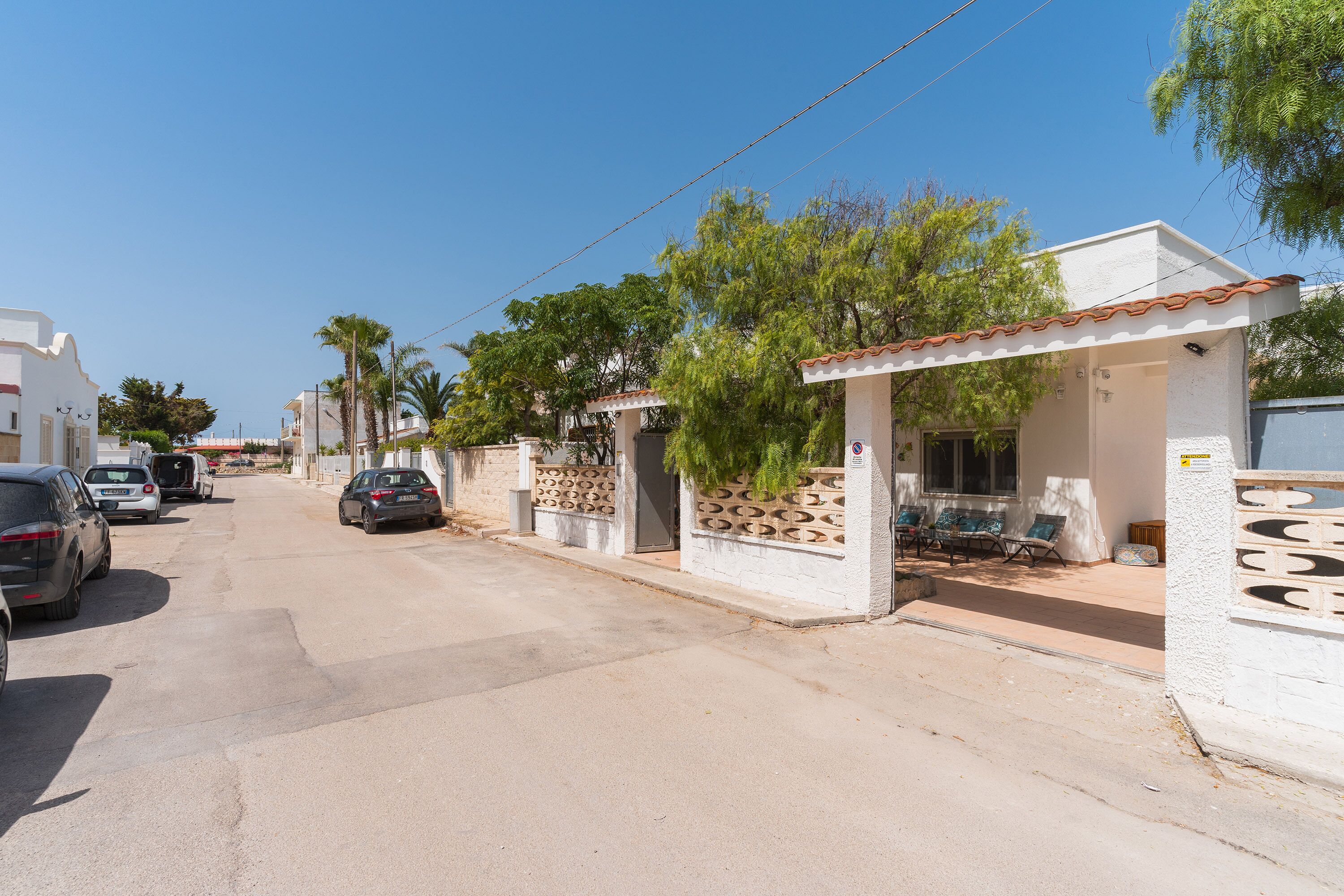 A residential street with parked cars, houses with tiled roofs, and palm trees.
