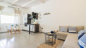 A modern living room with a sofa, a dining table with chairs, a kitchen area with cabinets and a countertop, and a small round table with a rug. - 2473 Villetta Seaside by Barbarhouse (Porto Cesareo)