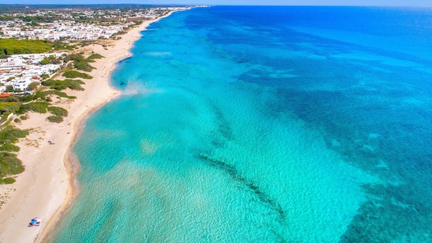 An aerial view of a beach with clear turquoise waters, a sandy shoreline, and a residential area with buildings and greenery.