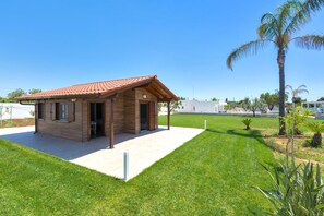 A small wooden house with a tiled roof, a covered patio, and a well-maintained lawn with palm trees.