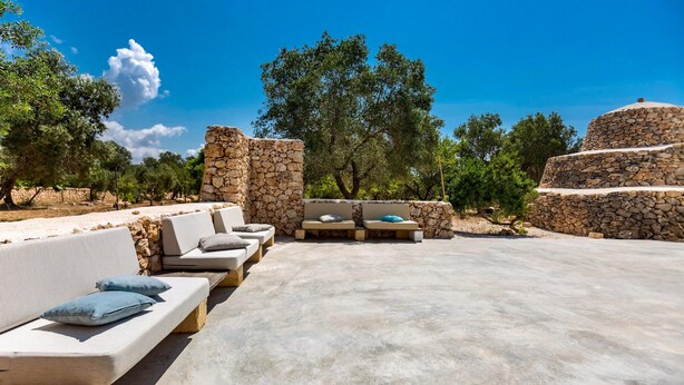 An outdoor seating area with stone walls and sofas, surrounded by trees and a clear sky. - 2670 Trullo Corni di Fiasco by Barbarhouse (Maruggio)