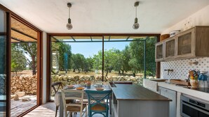A modern kitchen with a large island, wooden cabinets, and a view of a stone wall and trees. - 2670 Trullo Corni di Fiasco by Barbarhouse (Maruggio)