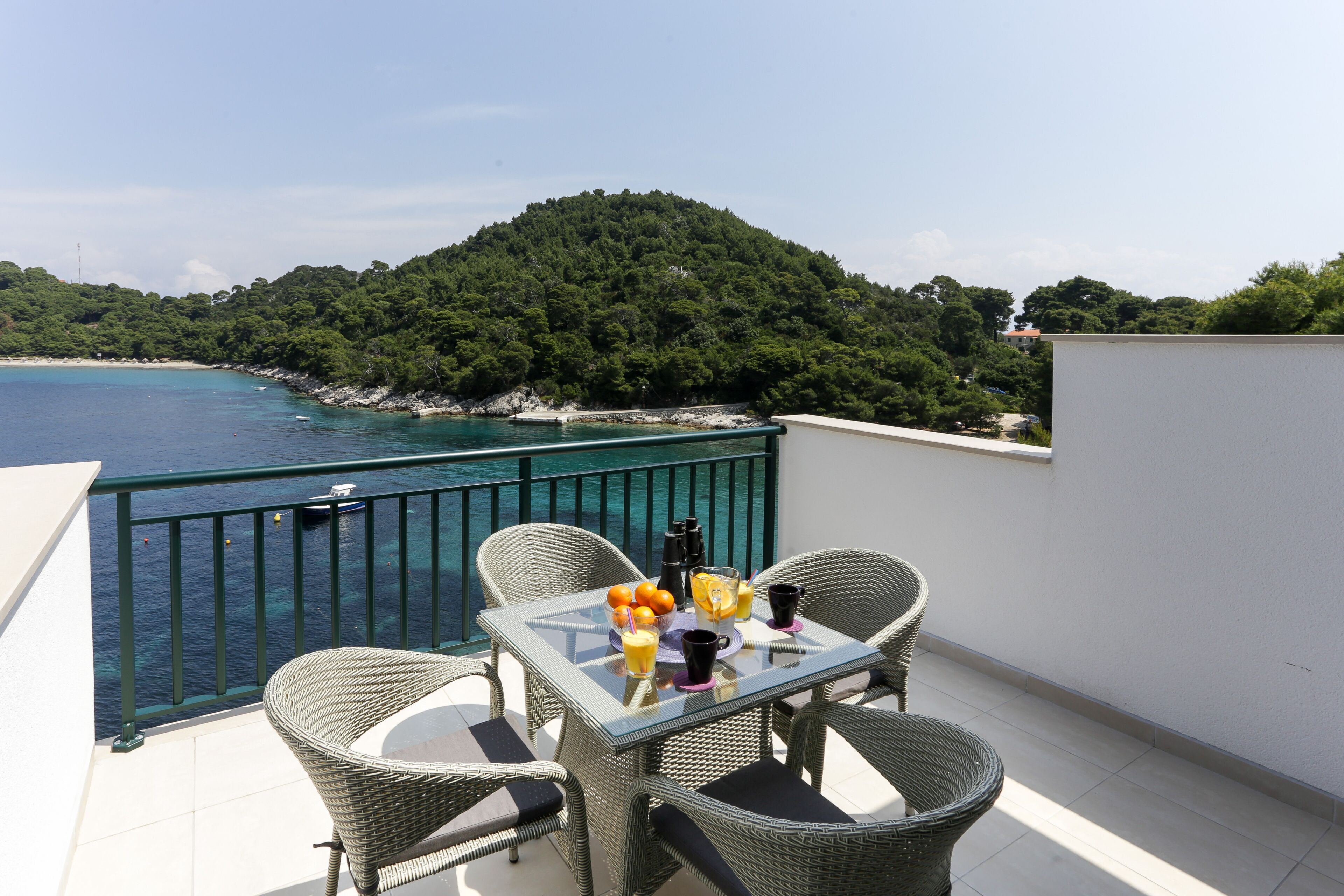 Un balcone con mobili in vimini, un tavolo con frutta e bevande, e vista sul mare e su un pendio boscoso.