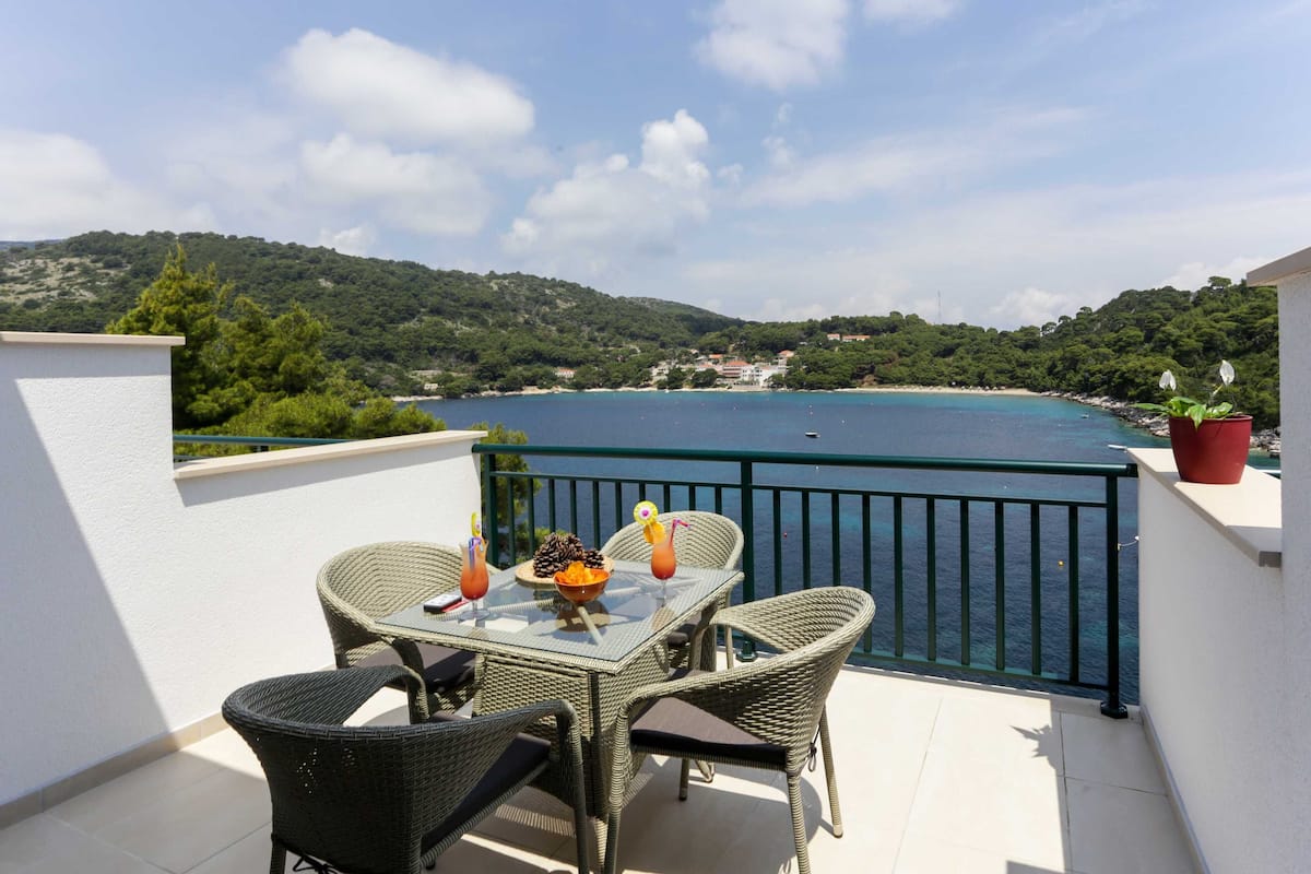 Ein Balkon mit Korbstühlen und einem Tisch, der Blick auf die Küste und einen klaren Himmel bietet.