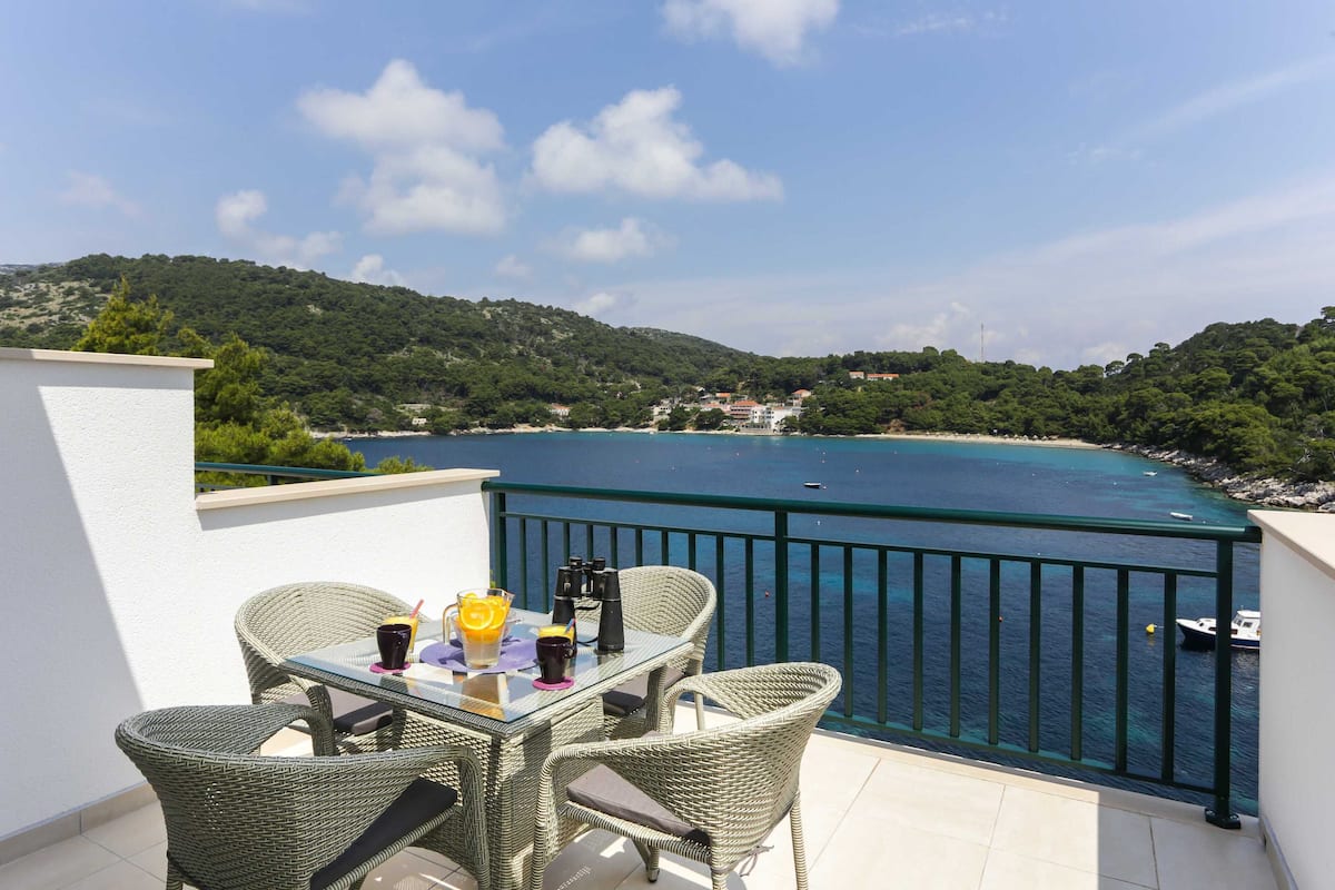 Ein Balkon mit Korbmöbeln und einem Tisch, mit Blick auf eine Küstenlandschaft mit klarem, blauem Wasser und einer hügeligen Umgebung.