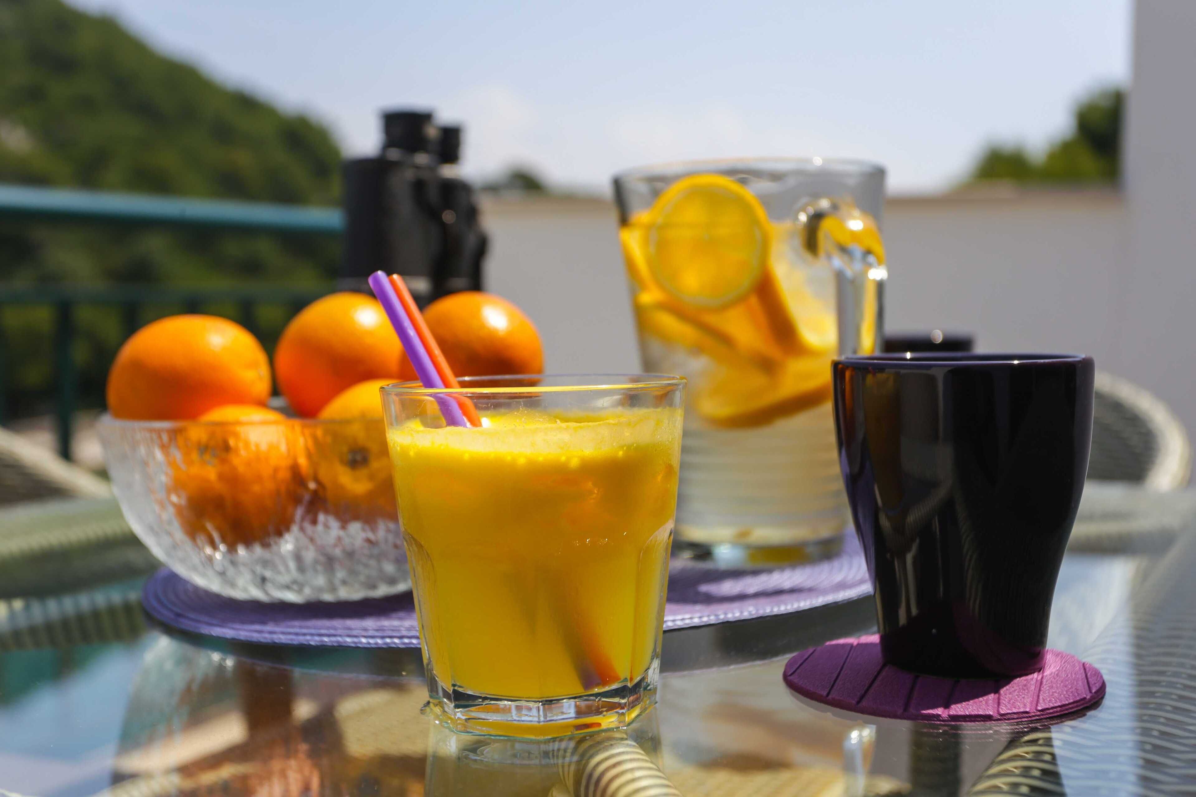 A glass of orange juice with a straw, a pitcher of orange juice, and a bowl of oranges on a table.
