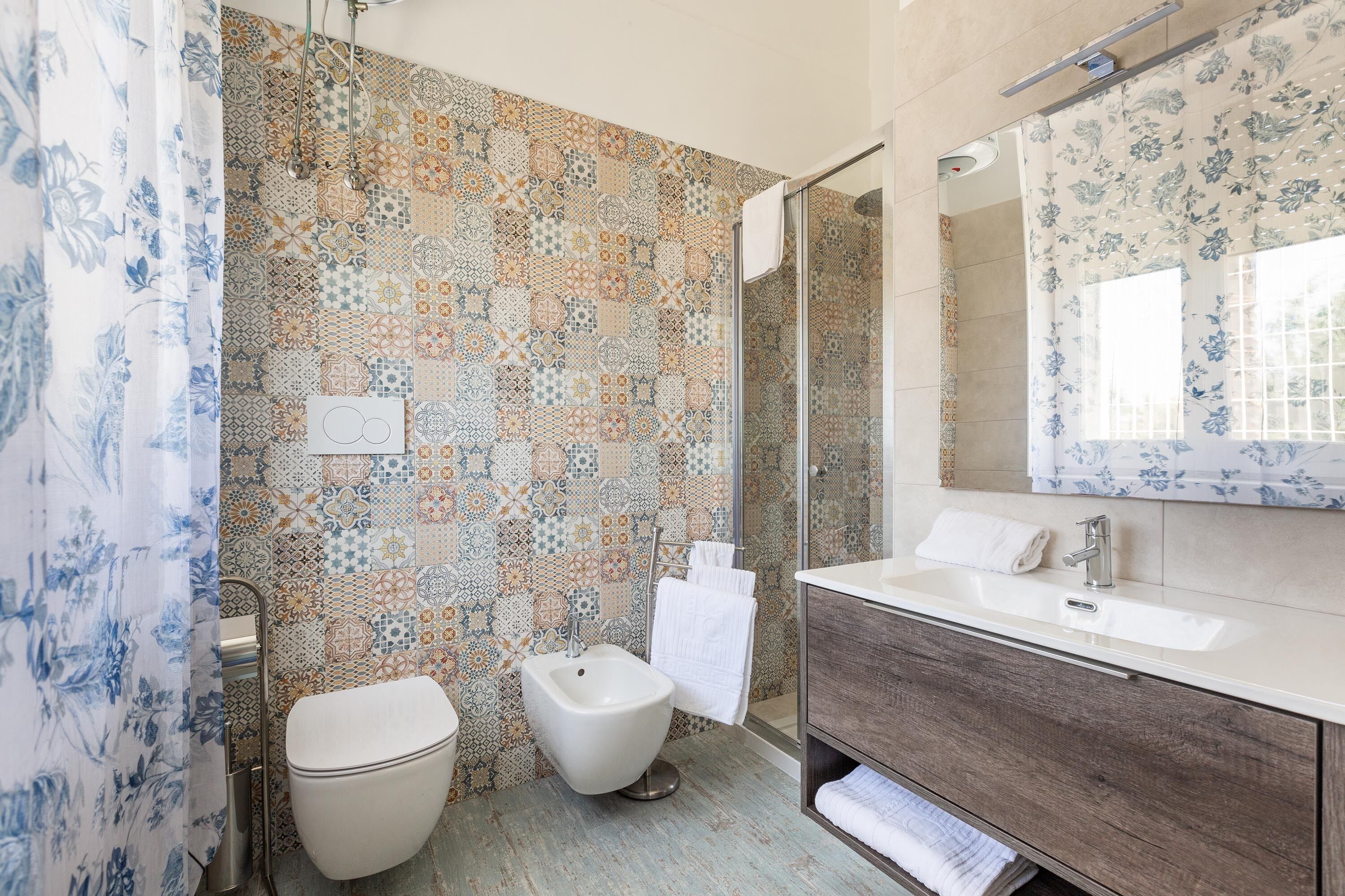 Et badeværelse med væg i fliser med mønster, toilet, bidet, brusebad og en kommode med spejl.
