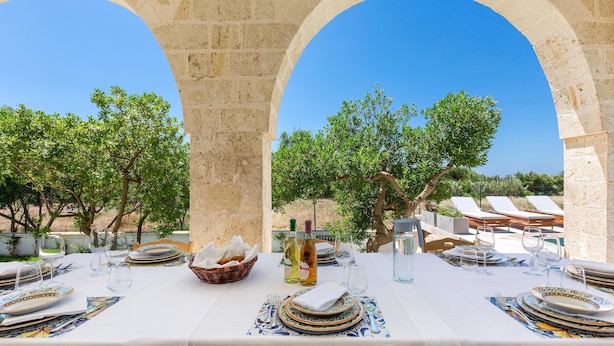 An outdoor dining area with a table set for two, under a stone archway, overlooking a pool area with lounge chairs.