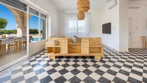 A modern living room with a checkered floor, a wooden sofa with cushions, a hanging light fixture, and a television on the wall.