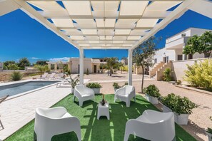 Ein überdachter Außenbereich mit Pool, weißen Liegestühlen und Kübelpflanzen.