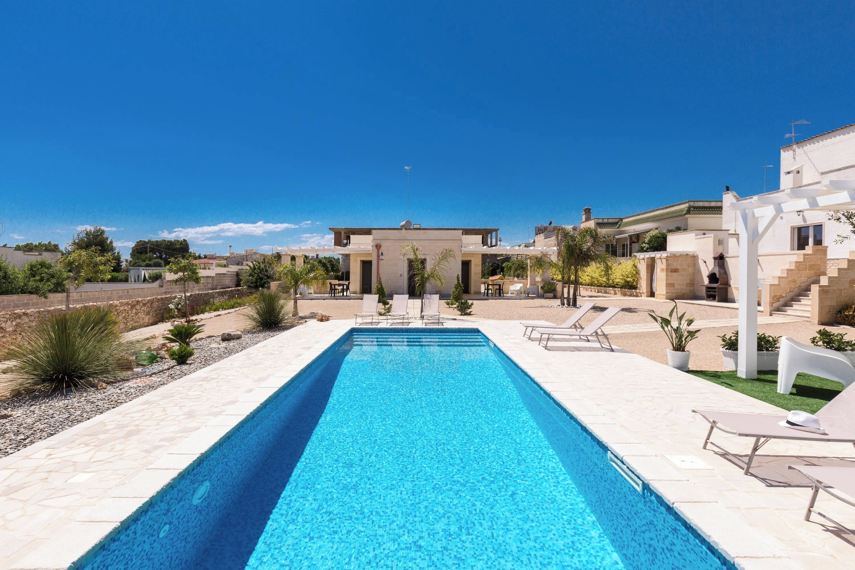 A rectangular swimming pool with a clear blue water surface, surrounded by a tiled deck. There are lounge chairs and a small garden area with plants and a white structure.