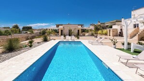 A rectangular swimming pool with a clear blue water surface, surrounded by a tiled deck. There are lounge chairs and a small garden area with plants and a white structure. - 2494 Villa Tina Maria by Perle di Puglia (Manduria)