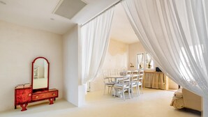Een slaapkamer met een bed, een kaptafel met spiegel, een eettafel met stoelen en dunne gordijnen.