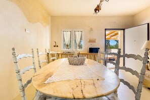 A dining area with a marble table, white lace tablecloth, and a wicker basket. There are white chairs with carved backs. A television is on a wooden table in the background.