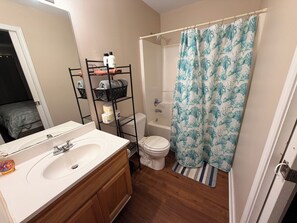 Un baño con lavamanos blanco, tocador de madera, estante negro con artículos de aseo, inodoro y cortina de ducha con estampado azul.