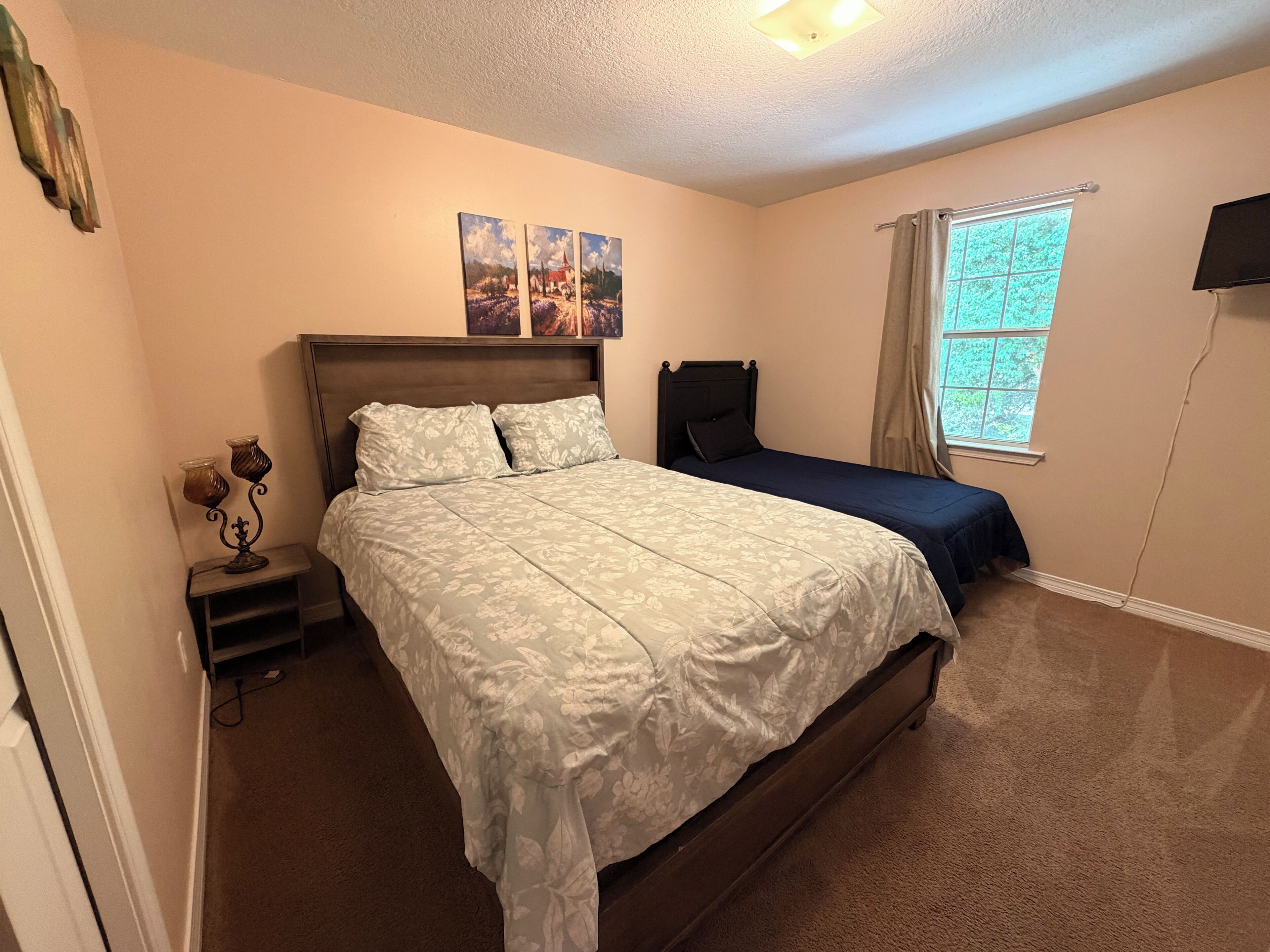 Une chambre avec un grand lit, un lit plus petit, une table de chevet avec une lampe, une fenêtre avec des rideaux et deux tableaux accrochés au mur.