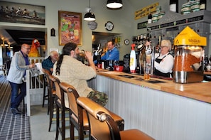 Een bar met houten barstoelen, een toonbank met diverse flessen en een grote bak met snacks.