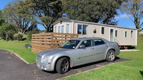Een zilverkleurige sedan geparkeerd voor een stacaravan met een houten terras en een schotelantenne.