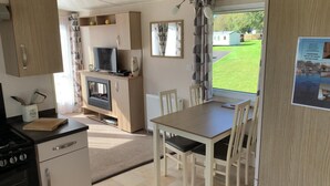 Cocina compacta con estufa, microondas y un pequeño televisor. Hay una mesa de comedor con sillas y una ventana que ofrece vistas a un césped verde.