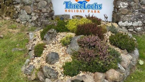 Un letrero de bienvenida para Three Lochs Holiday Park rodeado de un muro de piedra y follaje.