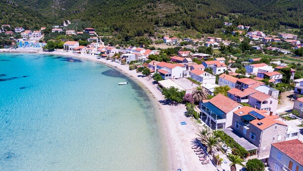 Un pueblo costero con casas a lo largo de la playa, aguas azules cristalinas y colinas verdes al fondo.
