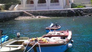 A coastal building with a red-tiled roof, a stone wall, and a clear blue water body with boats. - Guest House Hazdovac Rooms (Mljet)