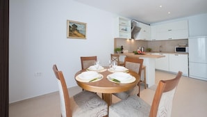 A dining area with a round wooden table set for four, surrounded by patterned chairs. A kitchen with white cabinets and a microwave is visible in the background.