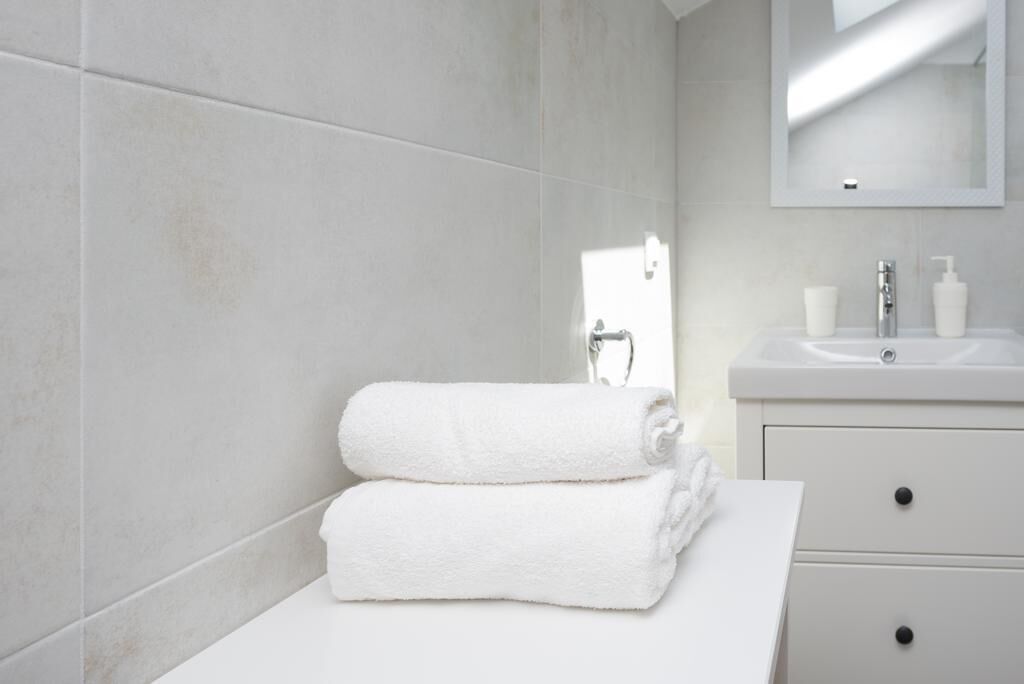 A bathroom with white tiles, a sink, a mirror, and two rolled towels on a white countertop.