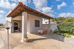 Uma pequena casa com telhado de telhas, um pátio com mesa e cadeiras, e um vaso de planta.