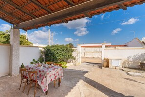 Pátio externo com mesa posta para refeições, cercado por cadeiras, sob um telhado de azulejos.