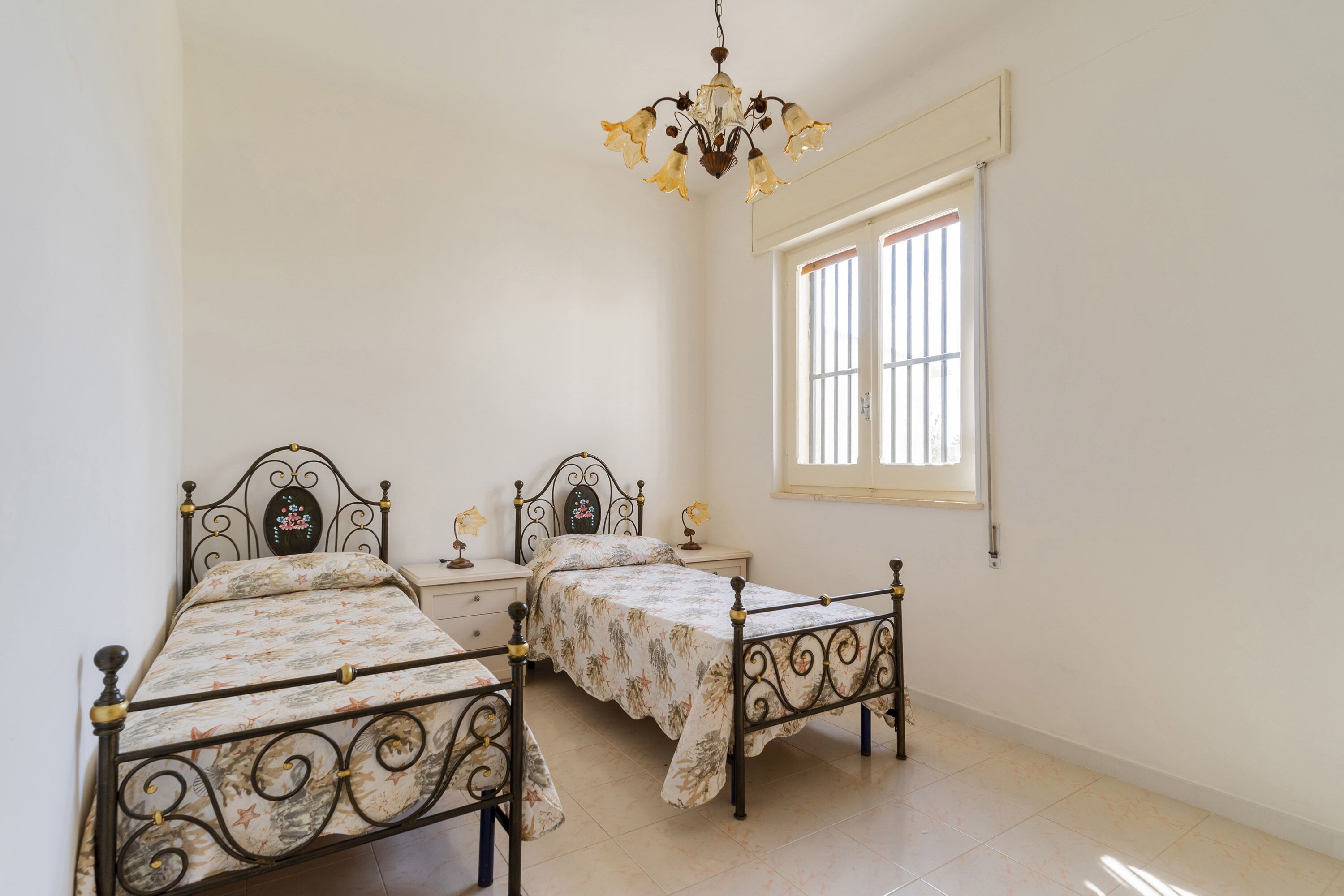 A bedroom with two metal beds, a chandelier, a window with bars, and a white wall.