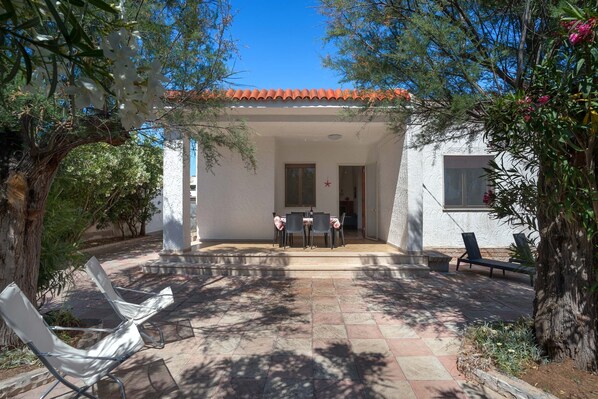 A covered patio with a table and chairs, surrounded by trees and a white building with a tiled roof.