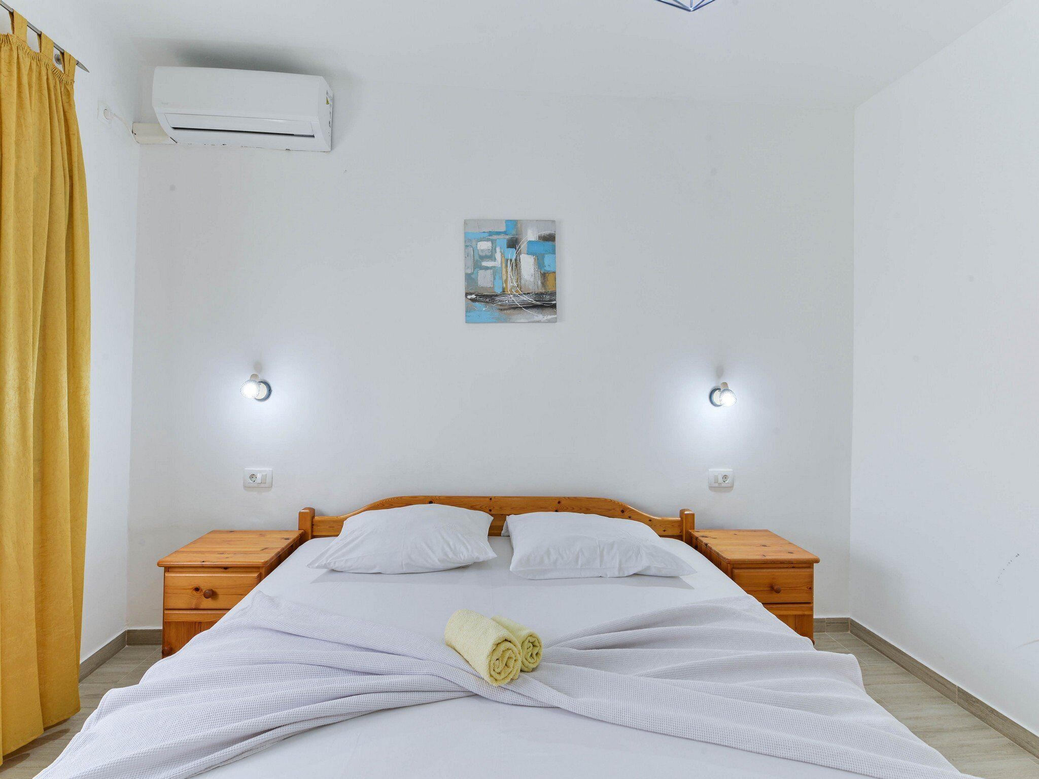 A hotel room with a bed, two bedside tables, an air conditioner, and a painting on the wall.