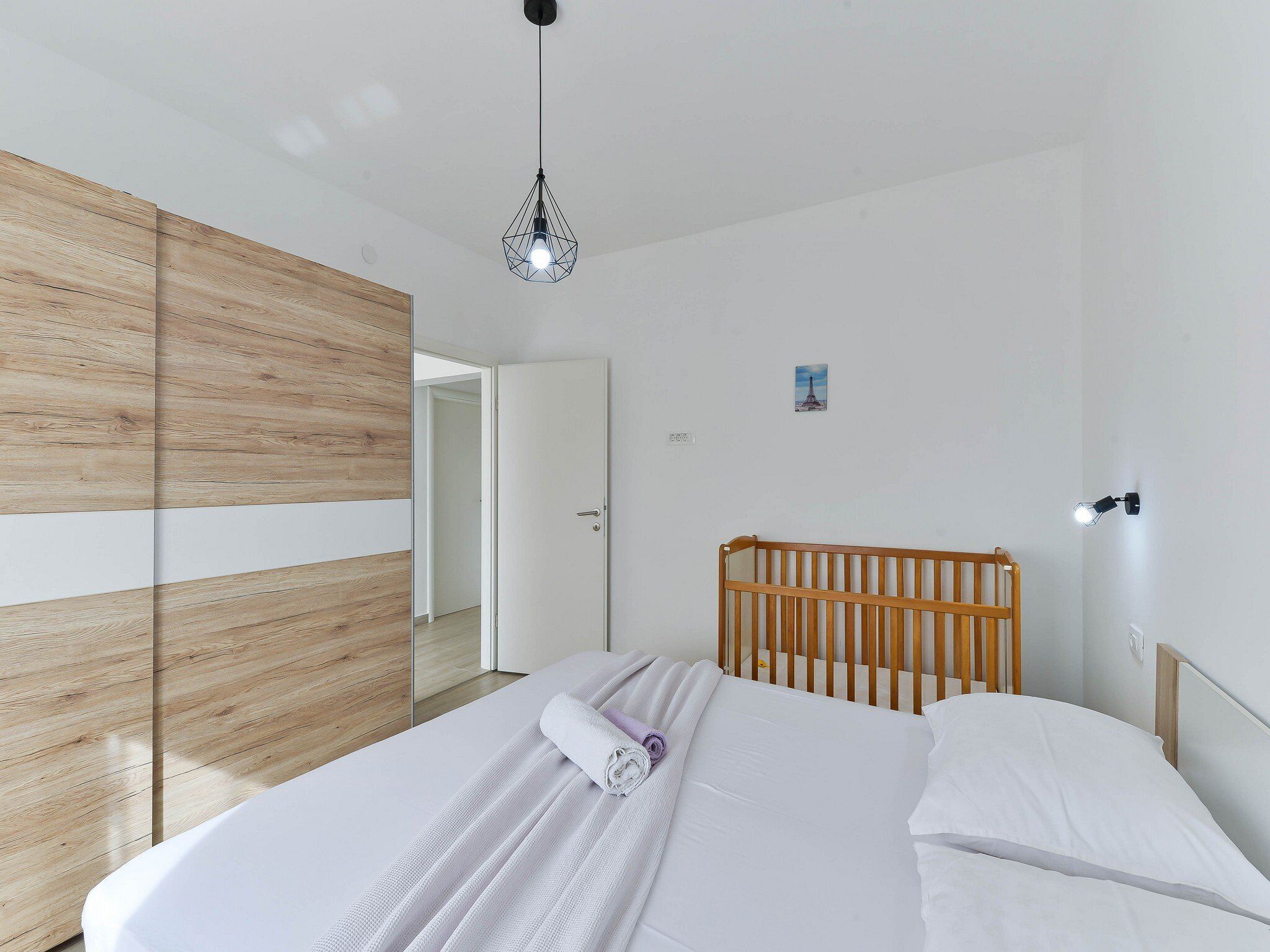 A modern bedroom with a wooden wardrobe, a bed with white linens, and a hanging light fixture.